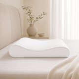 A white contoured memory foam pillow resting on a bed with a beige headboard and cream bedding, with a vase of green branches in the softly lit background.